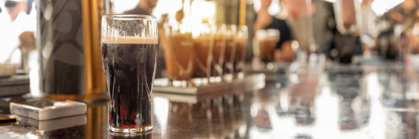 Glasses of stout beer on a bar counter, blurred people, panoramic pub header stock photo