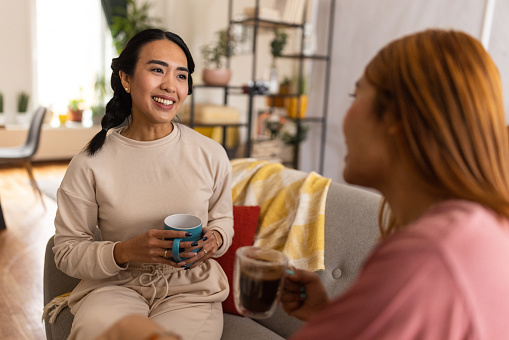 Two Asian female friends drinking coffee together in the living room of their house