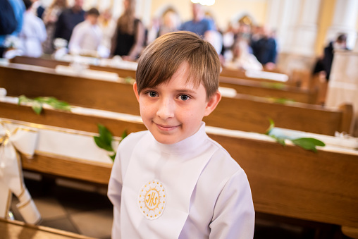 the boy holds a burning candle in his hands, stands in white clothes in the temple before the children's mass