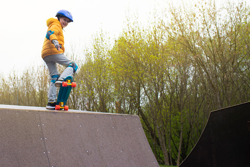 a boy in a yellow jacket, with blue protection on his arms and legs, with a purple helmet with a plastic skateboard in a skate park in cold weather