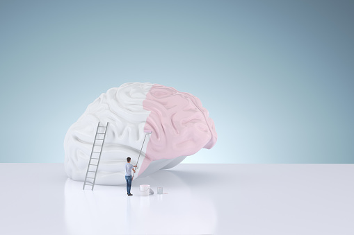 The symbolic human brain being painted pink by a human for positive emotions on a light blue background with copy space.  Digitally generated image.