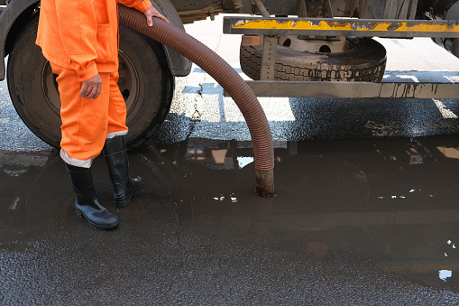 worker pumps out water with a special pump. cleaning of drains on the street. a man in an orange uniform and black rubber boots close-up.