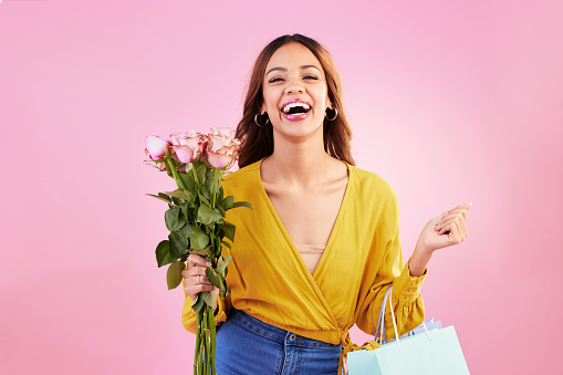 Happy, shopping bags and flowers with woman in studio for retail, birthday and spring. Event, party and celebration with female customer and roses on pink background for sale, discount and romance