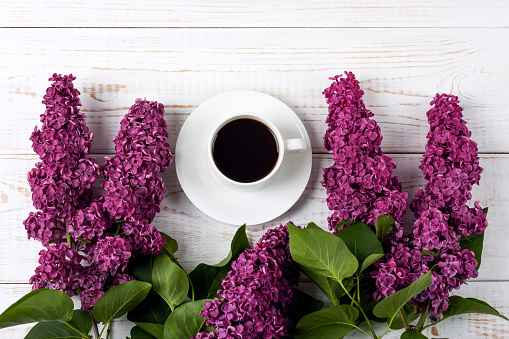 Cup of fresh black coffee and branch of blooming purple lilac with green leaves on white wooden background. Coffee break. Spring flowers. Flat lay, top view, copy space.