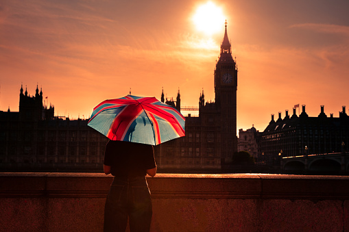 Color image depicting an unrecognisable woman holding an umbrella with the design of the British union flag on it. The woman is sitting on a bench overlooking the river Thames, the Houses of Parliament and Big Ben. A dramatic golden sunset is developing and the woman is looking at this iconic view.