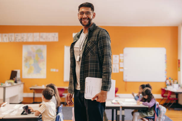 istock Happy elementary school teacher standing in a classroom, with children in the background 1495356247