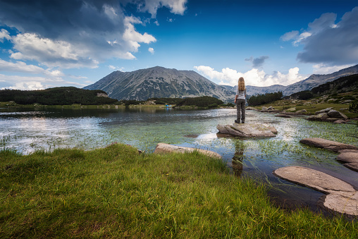 Rear view of a blonde woman enjoys the view of a beautiful lake and mountain peak behind, Todorka peak and Muratovo lake in Pirin mountains, Bulgaria