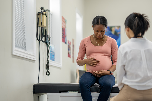 A young pregnant woman sit up on an exam table in her doctors office during a routine prenatal check-up.  She is dressed casually and her doctor is seated across from her as the two talk.