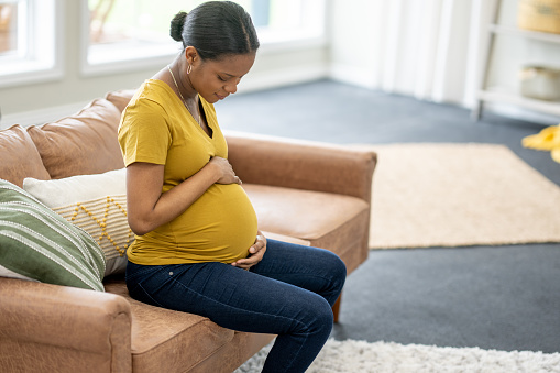 A young pregnant woman of African decent sits on a sofa in the comfort of her own home as she cradles her belly with her hands.  She is dressed casually as she looks down at her belly with anticipation.
