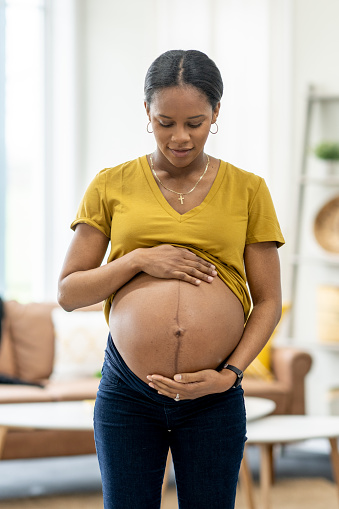 A young pregnant woman of African decent, stands in the comfort of her own home as she poses for a portrait.  She has her shirt over her belly as she cradles it with her hands and gazes down at her growing baby.