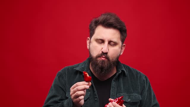 Bearded man eating red hot chilli pepper against red studio background. Spicy taste. Lover of hot, spicy food