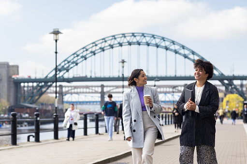 Two businesswomen walking together along the quayside and engaging in business-relate conversation. One of them is carrying a takeaway coffee and the other is carrying a diary under her arm. They are dressed in smart casual work clothing.