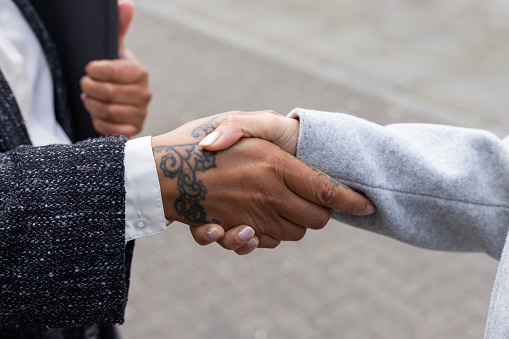 A close up unrecognisable shot of two business women shaking hands after securing a business deal together. They are both wearing smart casual work clothing. One of the women has a tattoo on her hand.