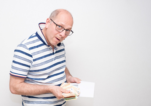 A 50-year-old Caucasian man on a gray background in a light shirt takes out a pack of euro bills from an envelope and looks slyly into the lens. series of emotions.