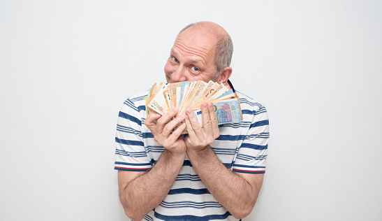 A Caucasian man 50 years old on a gray background in a light shirt holds a pack of euro bills in his hands and looks slyly into the camera. Series of emotions, sly face.