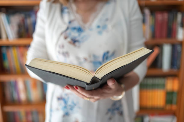 istock Woman standing with an open book while reading with a large bookcase behind her. 1495390352