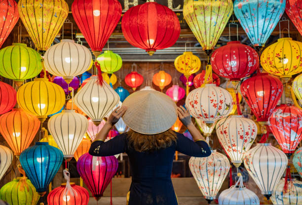 istock Back view anonymous woman in dress touching Asian conical hat on head while standing near colorful lanterns hanging on ceiling 1495393777