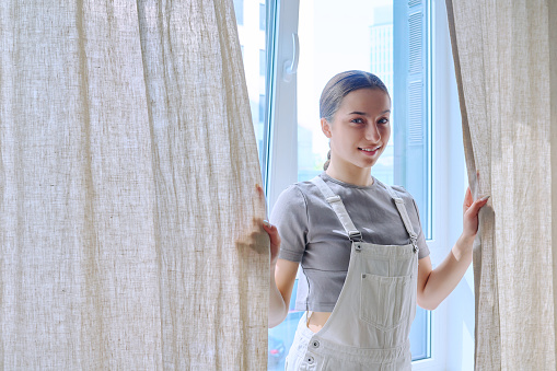 Smiling teenage girl looking at camera standing near window with curtains. 15,16 years old age, services for teens beauty health high school copy space, curtains place for advertising information text