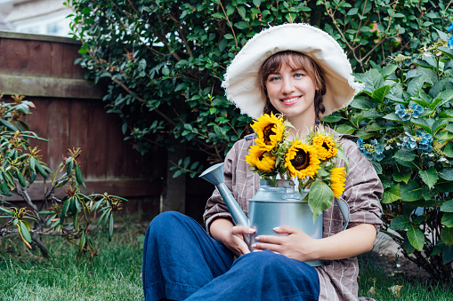 Portrait of smiling young female farmer woman holding watering can with fresh sunflowes bouquet while sitting in the green garden background. Cottagecore lifestyle. Selective focus, copy space
