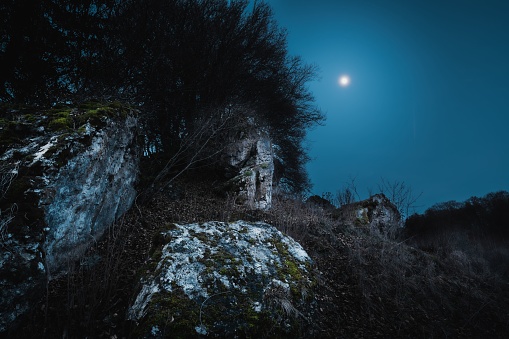 A full moon illuminates a rocky cliff face with jagged rocks silhouetted against the night sky
