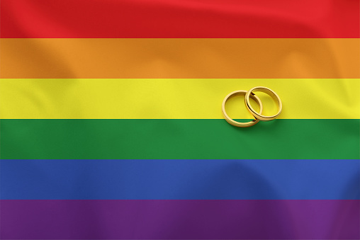 Conceptual detail of wedding between people of the lgbt collective with gold rings on textured flag with rainbow colors. Top view.