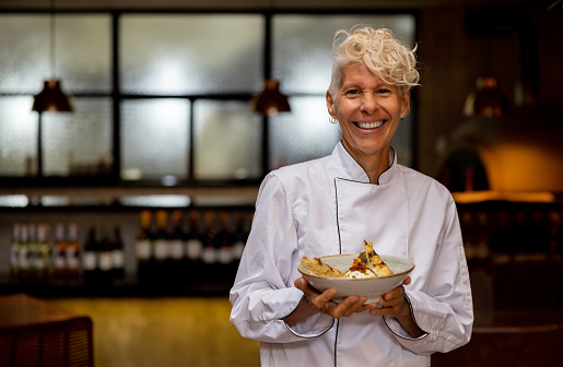 Portrait of a happy Latin American female chef holding a plate at a fine dining restaurant and looking at the camera smiling
