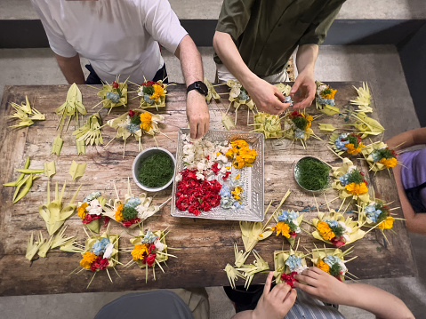 Overhead view of a multiracial family learning how to make traditional Balinese gratitude offerings in a Balinese culture class.  Using sustainable coconut leaves, cut pandan leaves and locally grown flowers.  Ubud, Bali, Indonesia