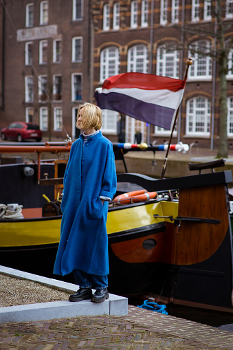 A woman in a stylish blue coat on a canal with ships and a waving flag of France. Feelings of passion for travel and adventure. Residential area in the Netherlands.
