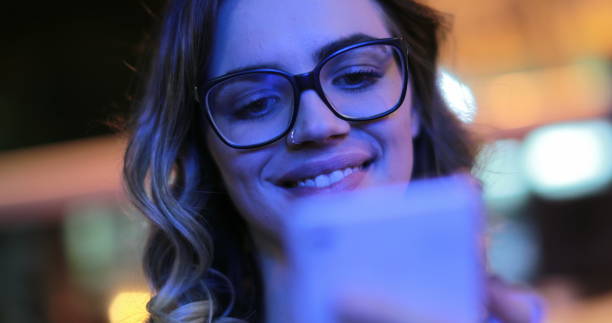 Girl wearing glasses checking cellphone at night in city atmosphere. Young woman smiling holding smartphone in the evening stock photo
