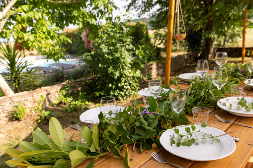 A medium close up side view of a table set for dinner. It has been beautifully decorated with lush foliage and white crockery and silver cutlery. It has been set by a French villas host.