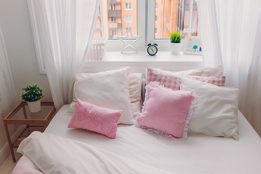 Horizontal shot of bed with white bedclothes, soft pillows, window with green plant, alarm clock and picture, no people. Coziness. Interior. Empty room with modern furniture