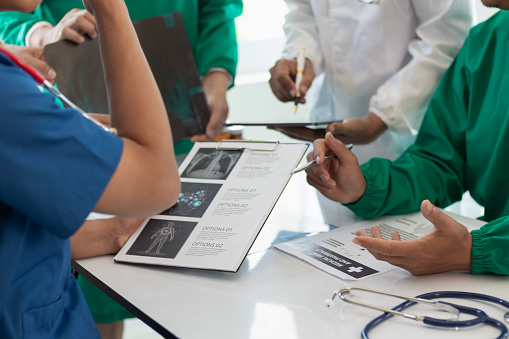 A Muslim woman wearing a hijab sits at a table while at a medical consultation. She has asthma and is learning to use an inhaler. A female doctor is watching and instructing the patient on how to use a puffer. The doctor's back is to the camera.