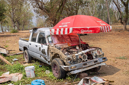 Luang Prabang, Laos - March 18th 2023: Wreck of a car where somebody set up a parasol to be able to harvest spare parts in shadow in a suburb to the former capital of Laos