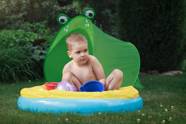 маленький мальчик сидит на корточках в надувном бассейне на лужайке во дворе, за зелеными палатками в форме лягушки - frog animal little boys child стоковые фото и изображения