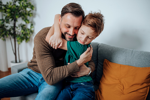 Experience the depth of a father's love as a mid adult man holds his son close on the sofa, cherishing the precious moments and nurturing a lifelong connection