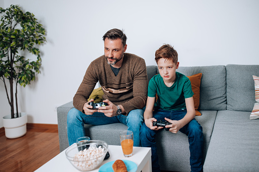 The mid adult man and his son share an incredible bonding experience as they dive into thrilling virtual adventures, comfortably seated on the sofa, fully immersed in the world of video games
