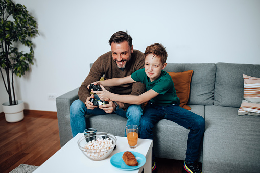 Witness the impressive skills and teamwork of the mid adult man and his son as they dominate the virtual gaming world, seamlessly coordinating their actions with precision, all from the comfort of their beloved sofa