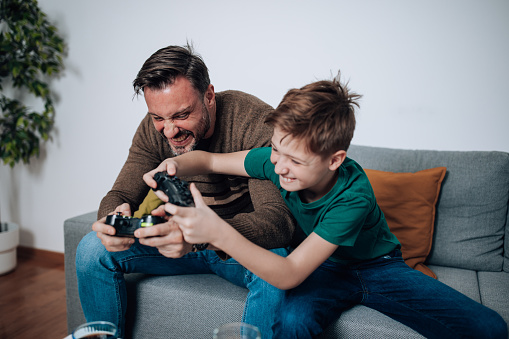 Beyond the pixels and screens, the mid adult man and his son strengthen their bond as they engage in meaningful conversations and shared experiences while indulging in their favorite video games on the sofa