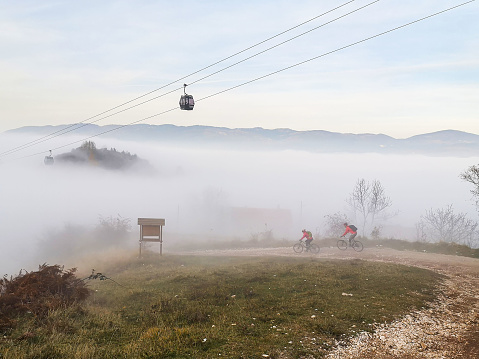 Cable car cabin above the city in the fog with a dirt road in the foreground with two cyclists