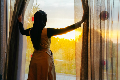 asian muslim woman looking throughout window at the sunset