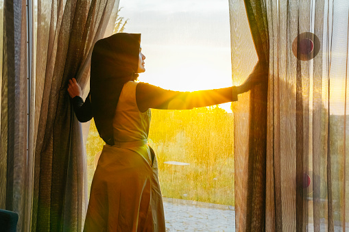 asian muslim woman looking throughout window at the sunset