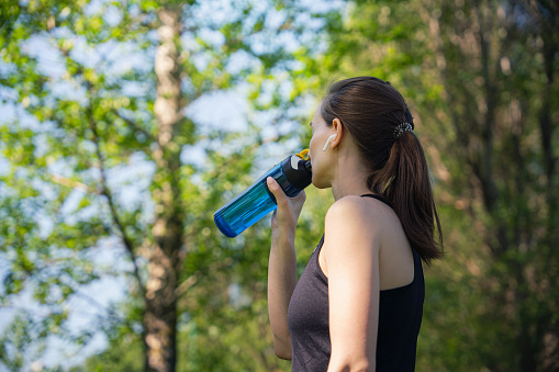Woman drinking water while jogging in the woods at sunny day