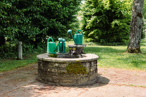 Group of green garden watering can in front of stone well. Romantic garden or parc, summertime secret garden scene.