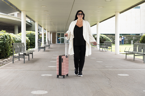 Mature woman in white cover-up leaving terminal and carrying her pink suitcase on wheels. Concept of business travel.Concept of business travel.