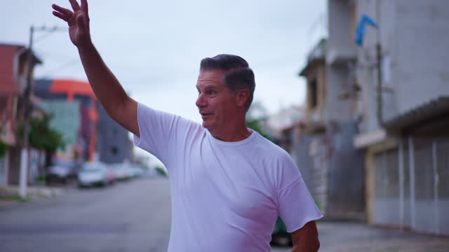 Happy older man waving hello to neighbors while walking in street. A middle-age caucasian male person walks forward toward camera while greeting to friends around him
