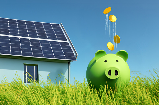 A green piggyback sits in the tall, juicy grass on a sunny day. Money rains from the sky into the piggybank. In the background, a solar powered detached house is visible. Energy saving concept