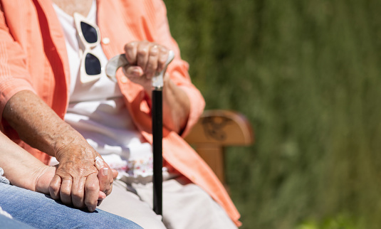 unrecognizable elderly woman with cane holding the hand of her daughter and caregiver.sitting together outdoors in summer on a bench.concept of support,help,comfort,advice,love and care.mental health