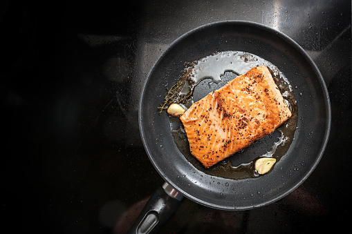 Fried salmon filet with with olive oil, herbs and garlic in a black frying pan on the stovetop, copy space, high angle view from above, selected focus, narrow depth of field