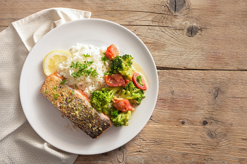 Crispy fried salmon filet with vegetables from broccoli and tomato, rice, parsley and lemon zest on a rustic wooden table, copy space, high angle view from above, selected focus