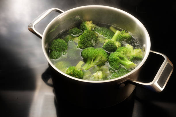 Cooking broccoli in boiling water in a stainless steel pot on the black cooktop in the kitchen, preparation for a healthy vegetable meal, copy space, selected focus, narrow depth of field stock photo